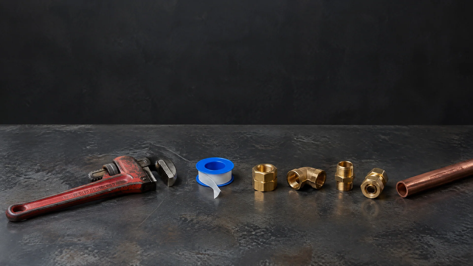 Pipe wrench, tape spool, gold-tone fittings and copper-colored tube in a row on a dark bench