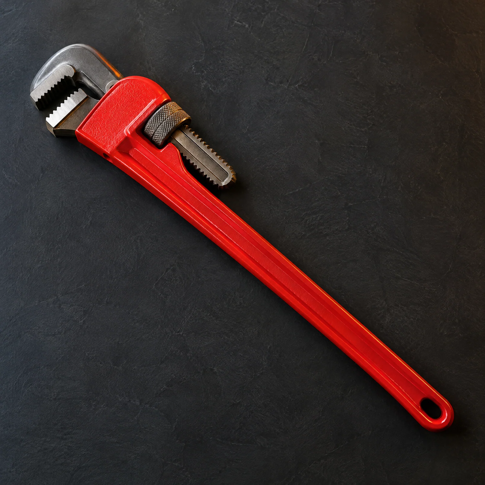 Red-handled pipe wrench with a toothed hook jaw on a dark slate surface